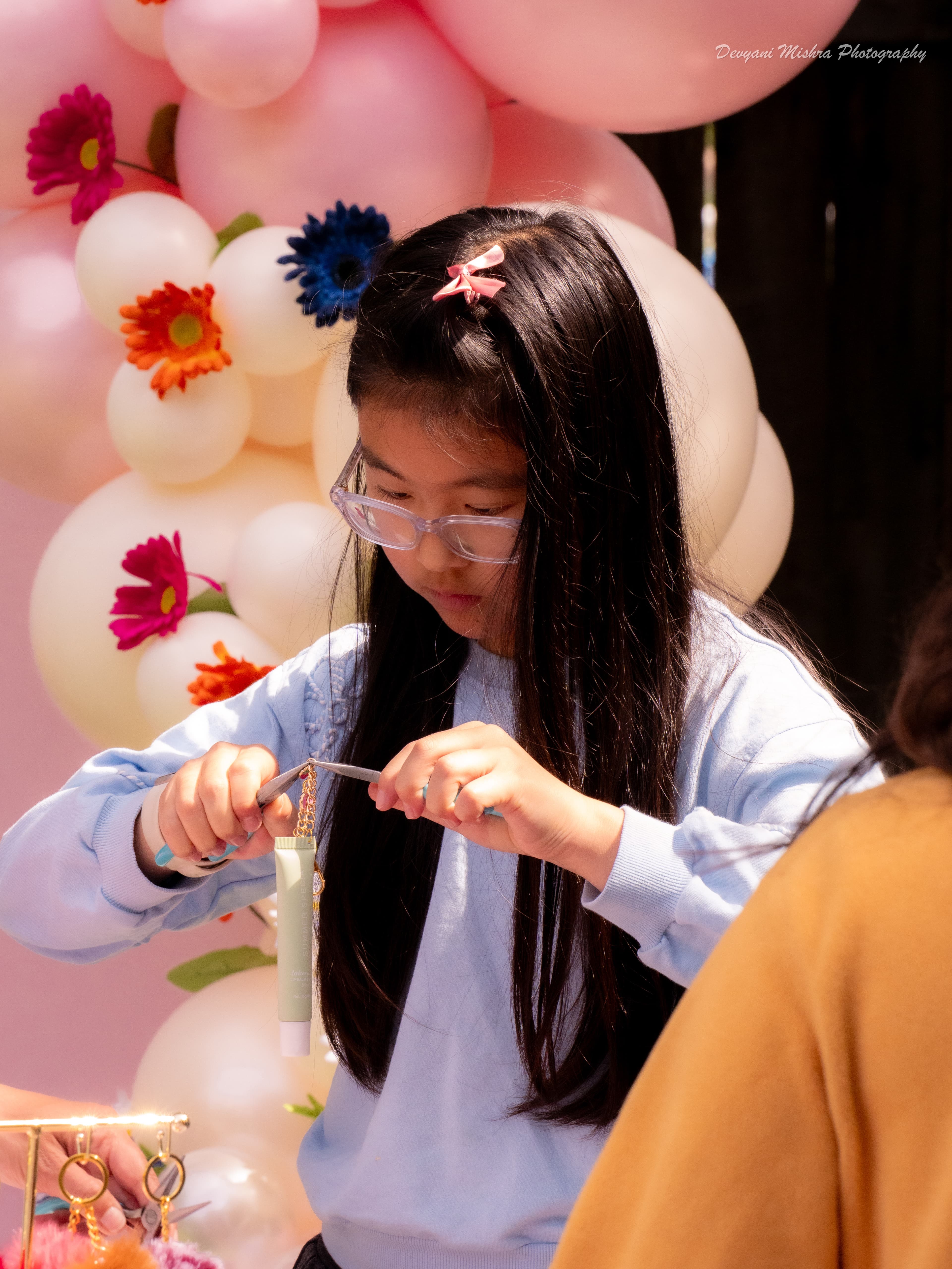 Girl threading a charm bracelet with pink floral balloon arch behind her
