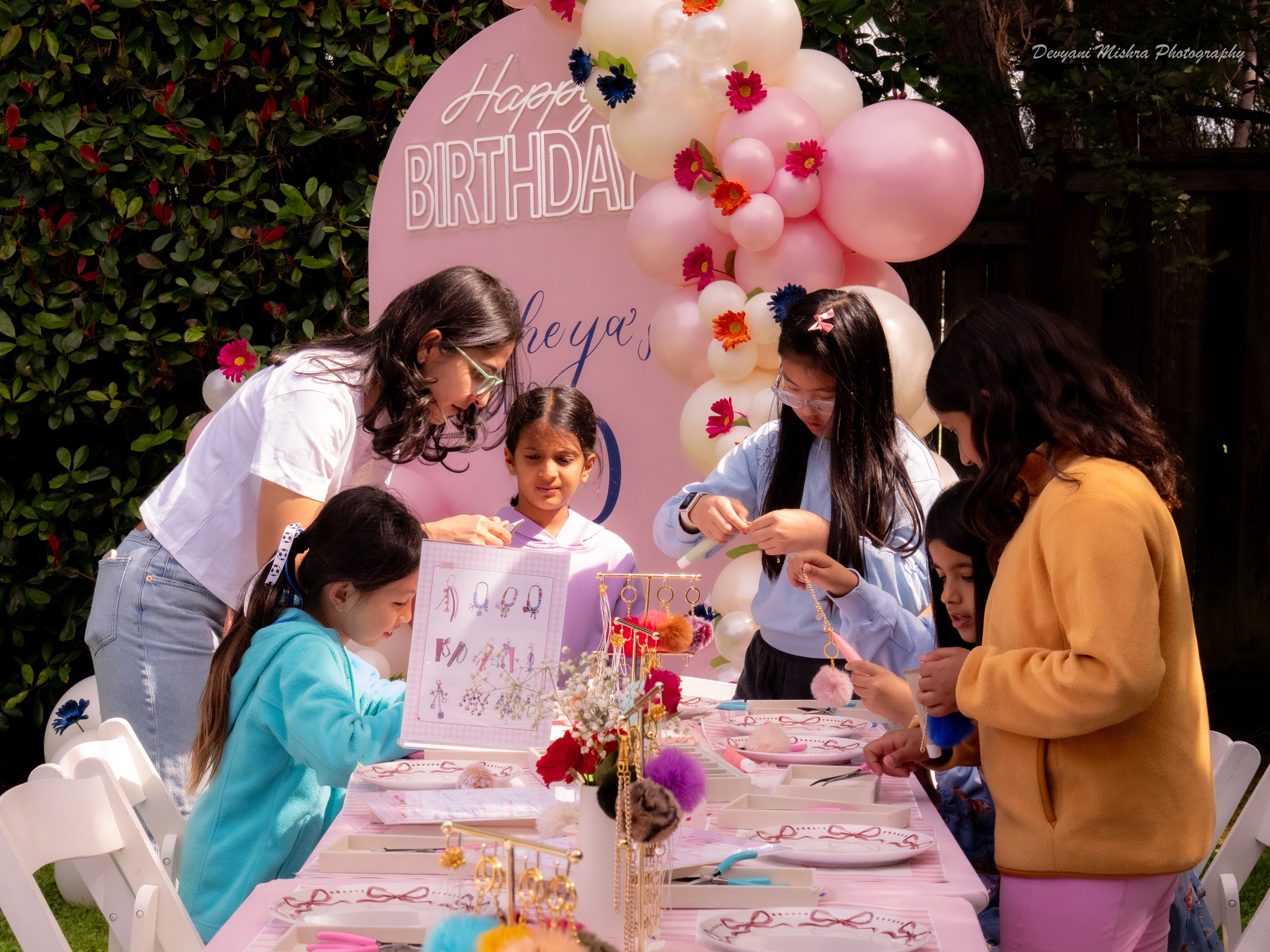 Wide shot of Rheya's birthday — kids at the Tag & Charm table with birthday backdrop and balloon arch