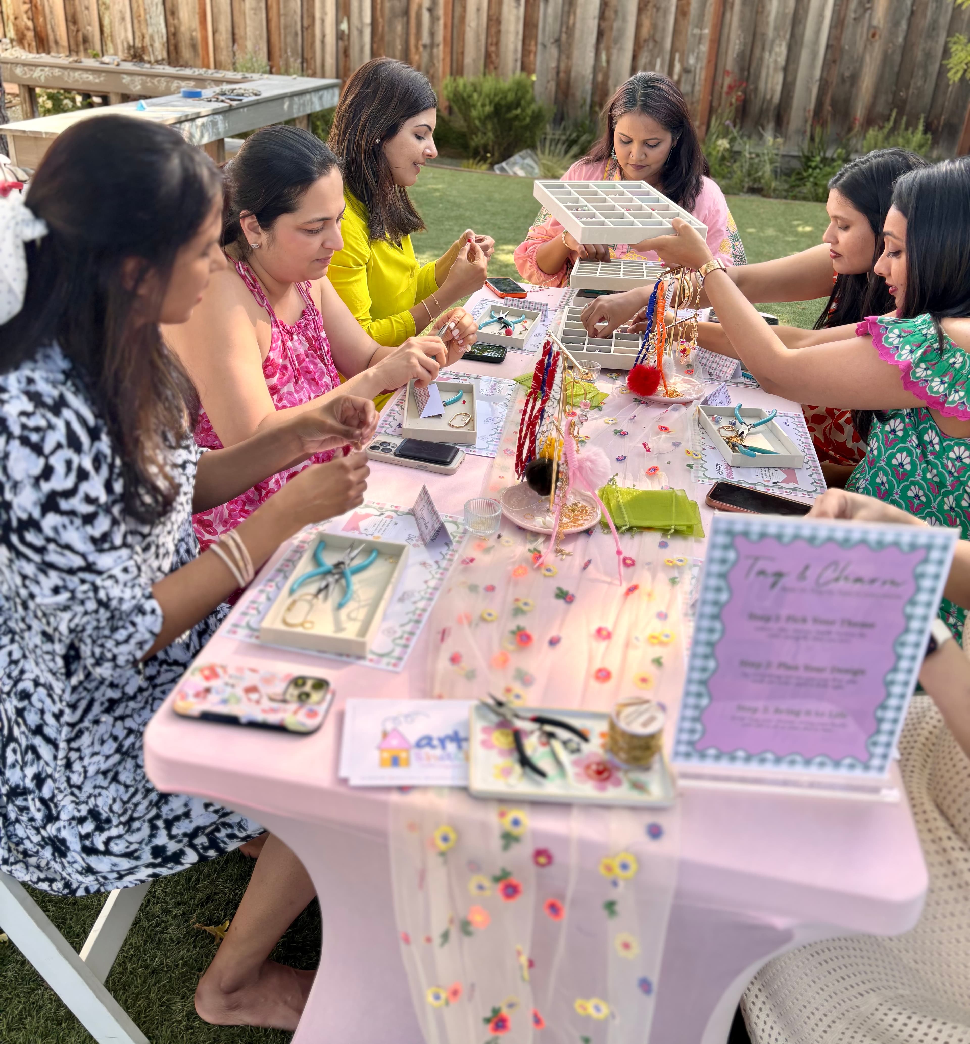 Six women in colorful outfits seated around the Tag & Charm table — wide shot